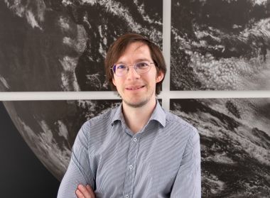 Dr. Mariano Mertens in front of a black-and-white satellite image of the Earth. He conducts research at the DLR Institute of Atmospheric Physics in Oberpfaffenhofen and is a junior research group leader of the IMPAC2T project from the funding measure "BMBF Junior Research Groups Global Change: Climate, Environment and Health".